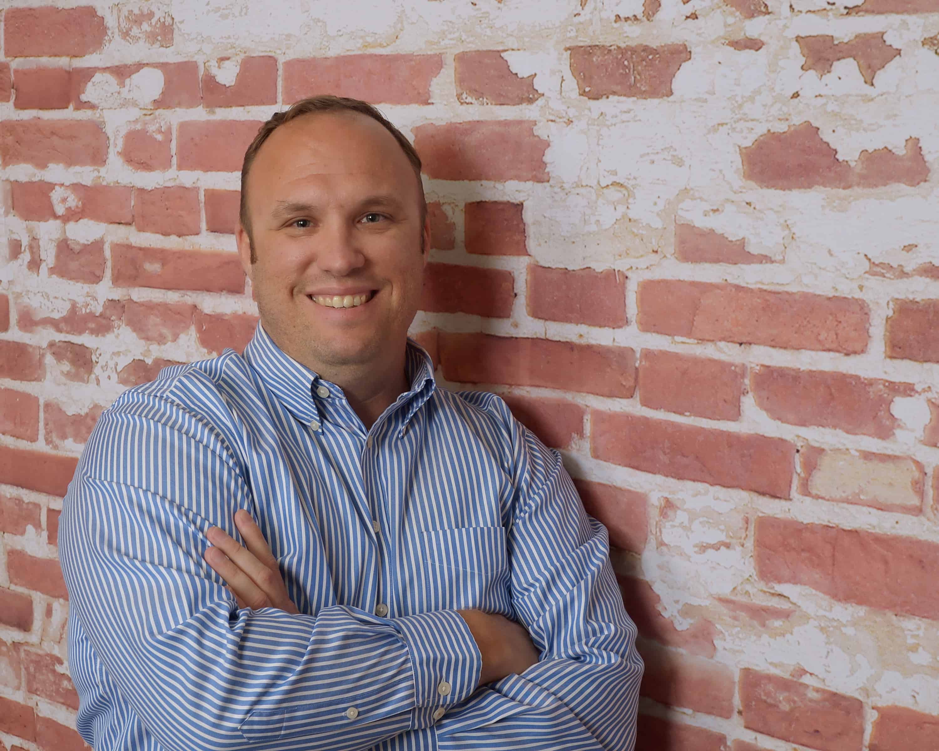 A smiling man in a blue striped shirt with arms crossed stands against a rustic red brick wall.