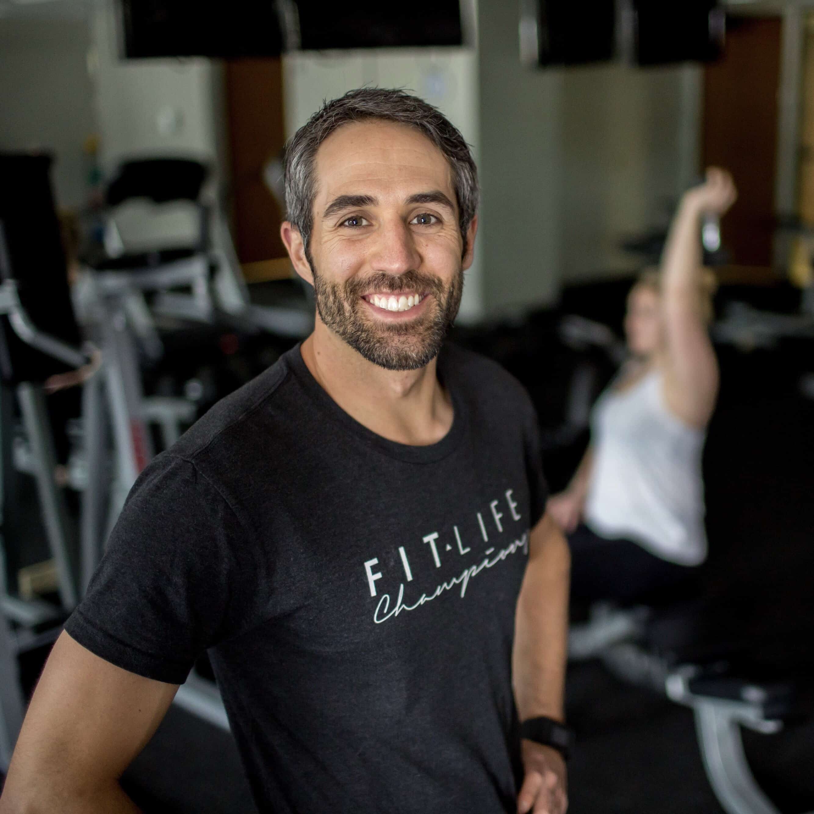 A smiling male fitness instructor named Dave Glaser, owner of Fit Life Champions, stands in a gym wearing a black t-shirt labeled "Fit Life Champion" with exercise equipment and a person stretching in the