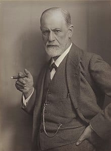 Sepia photo of an elderly man with a white beard, wearing a suit and holding a book, his hand on his hip, gazing sternly at the camera.